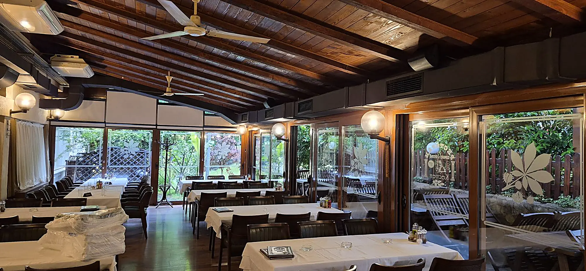 Zastakljena bašta restorana Kesten — drvena tavanica sa gredama, postavljeni stolovi i staklo koje gleda u zelenilo, sa ugraviranim listom kestena