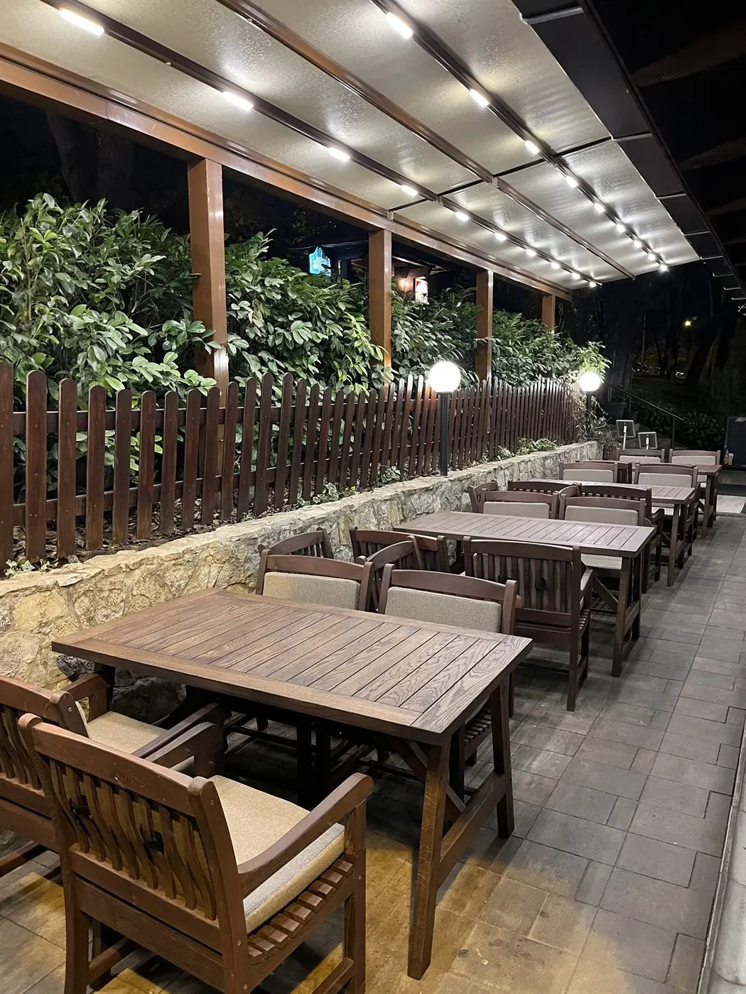 Terasa restorana Kesten uveče — osvetljena nadstrešnica, drveni stolovi uz kamenu ogradu i zelenilo iza ograde